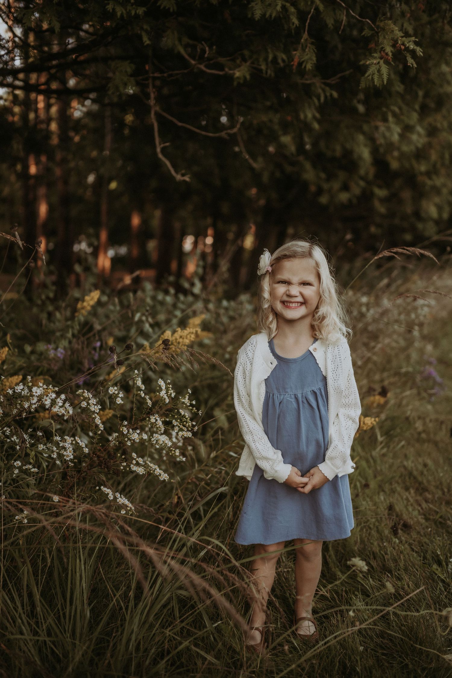 Young child in a blue dress and white cardigan standing in a wooded area with wildflowers.