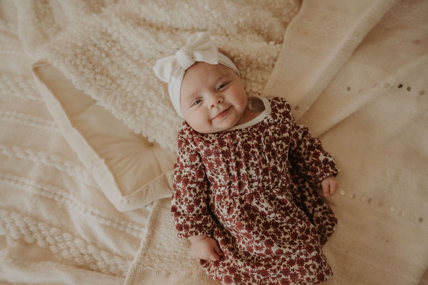 Baby wearing floral dress and white bow smiling on cream bedding.