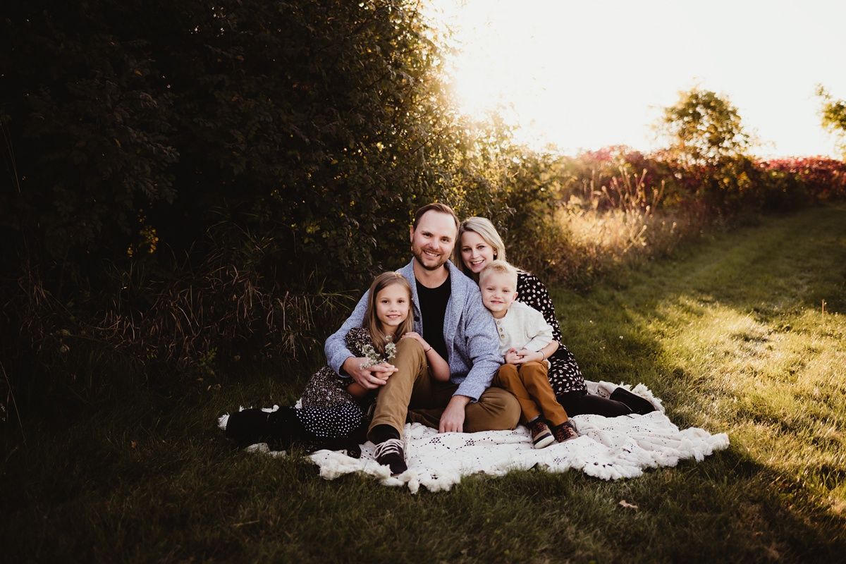 A family sits together on a white blanket during sunset in a grassy field.