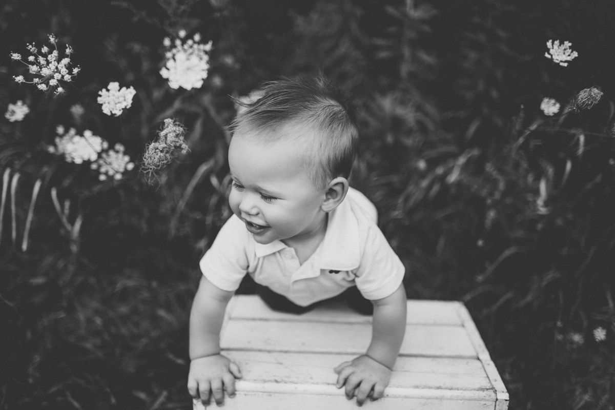 Black and white portrait of baby peering over wooden crate outdoors with flowers.