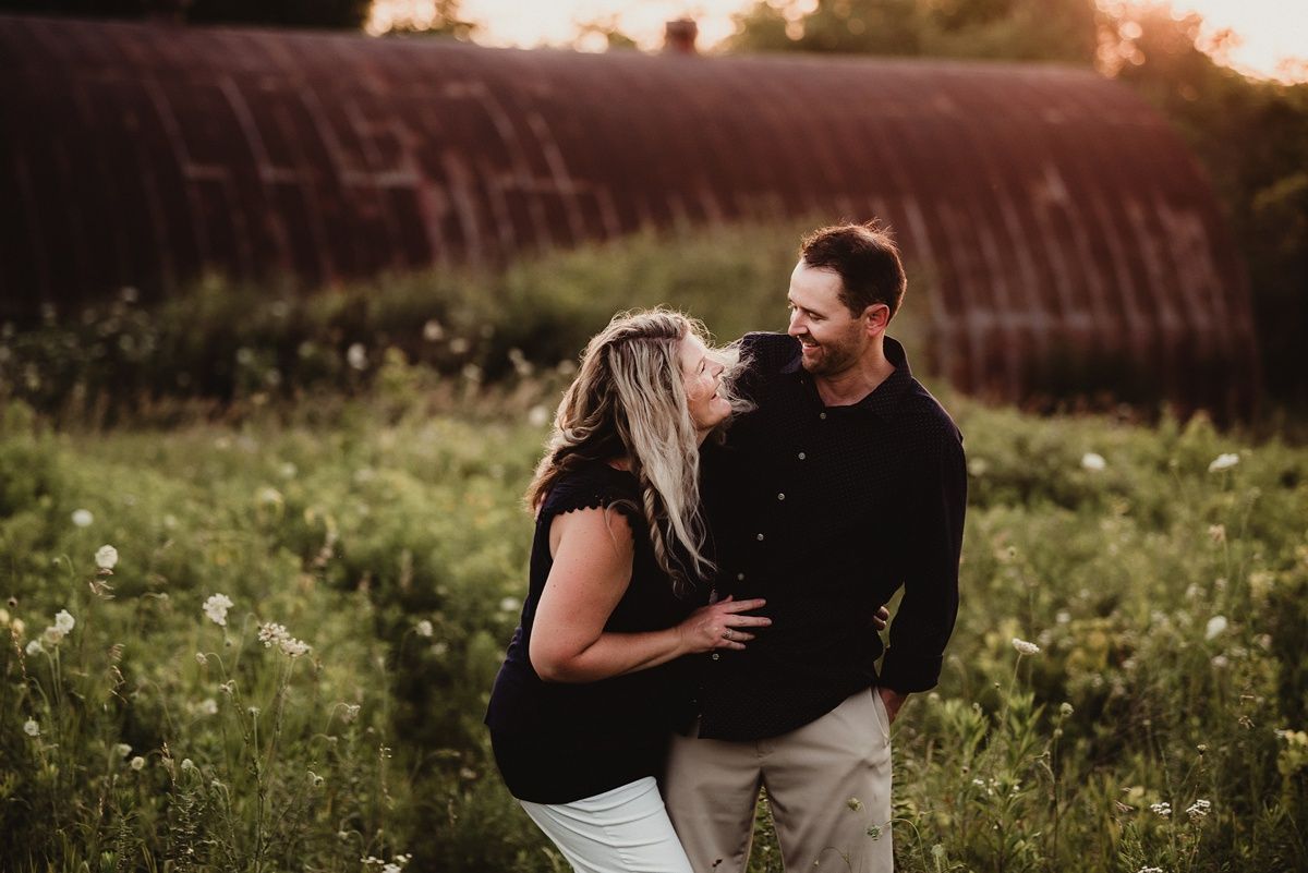 A couple shares an intimate moment in a golden field at sunset with a rustic barn behind them.