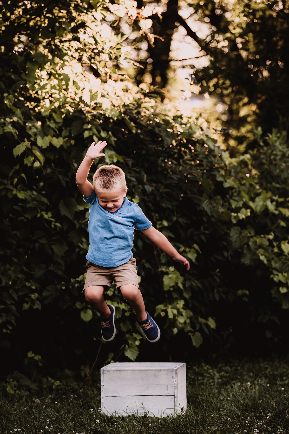 Young child joyfully jumping off white wooden crate in garden setting.