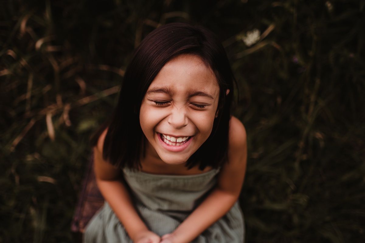 Joyful candid portrait of a young girl in a gray dress smiling brightly outdoors.