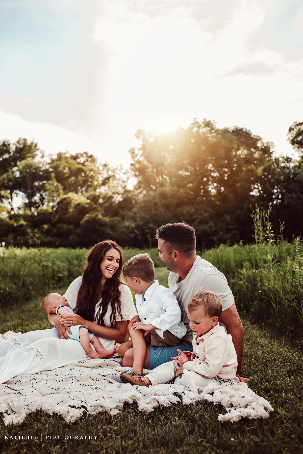 Family sitting together on white blanket in sunlit meadow during outdoor photo session.
