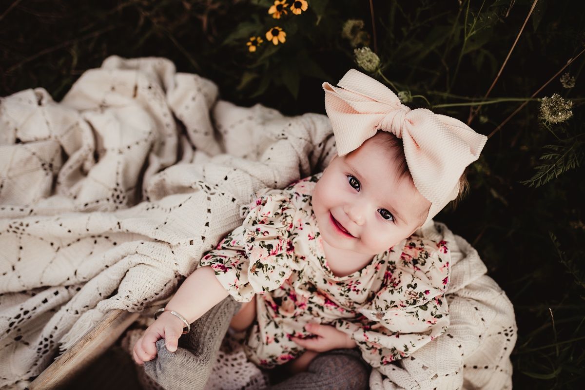 Baby in floral dress and large bow smiles while laying on cream blanket outdoors.