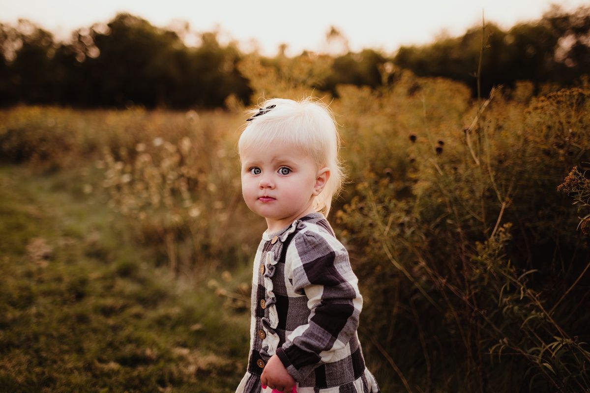 Young child in plaid dress stands thoughtfully among golden wildflowers at sunset.