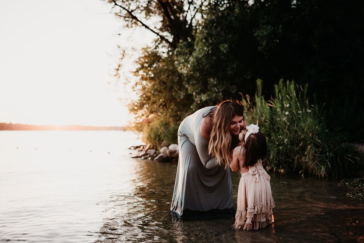 Tender lakeside moment between parent and child standing in shallow water at sunset.