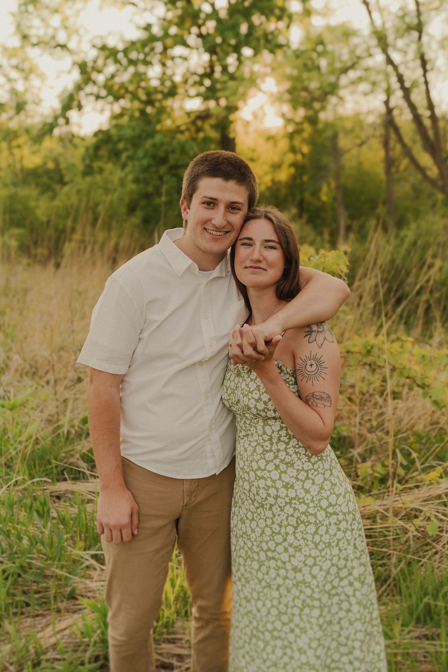 A casual outdoor portrait of a couple in coordinating outfits standing in tall grass at sunset.