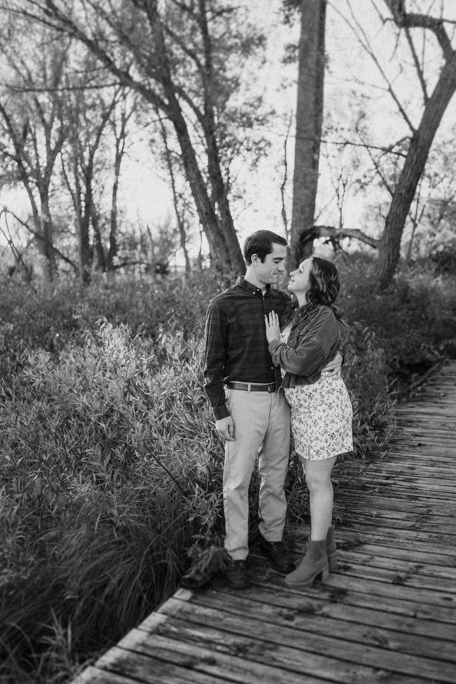 A romantic black and white photo of a couple standing on a wooden boardwalk surrounded by trees.