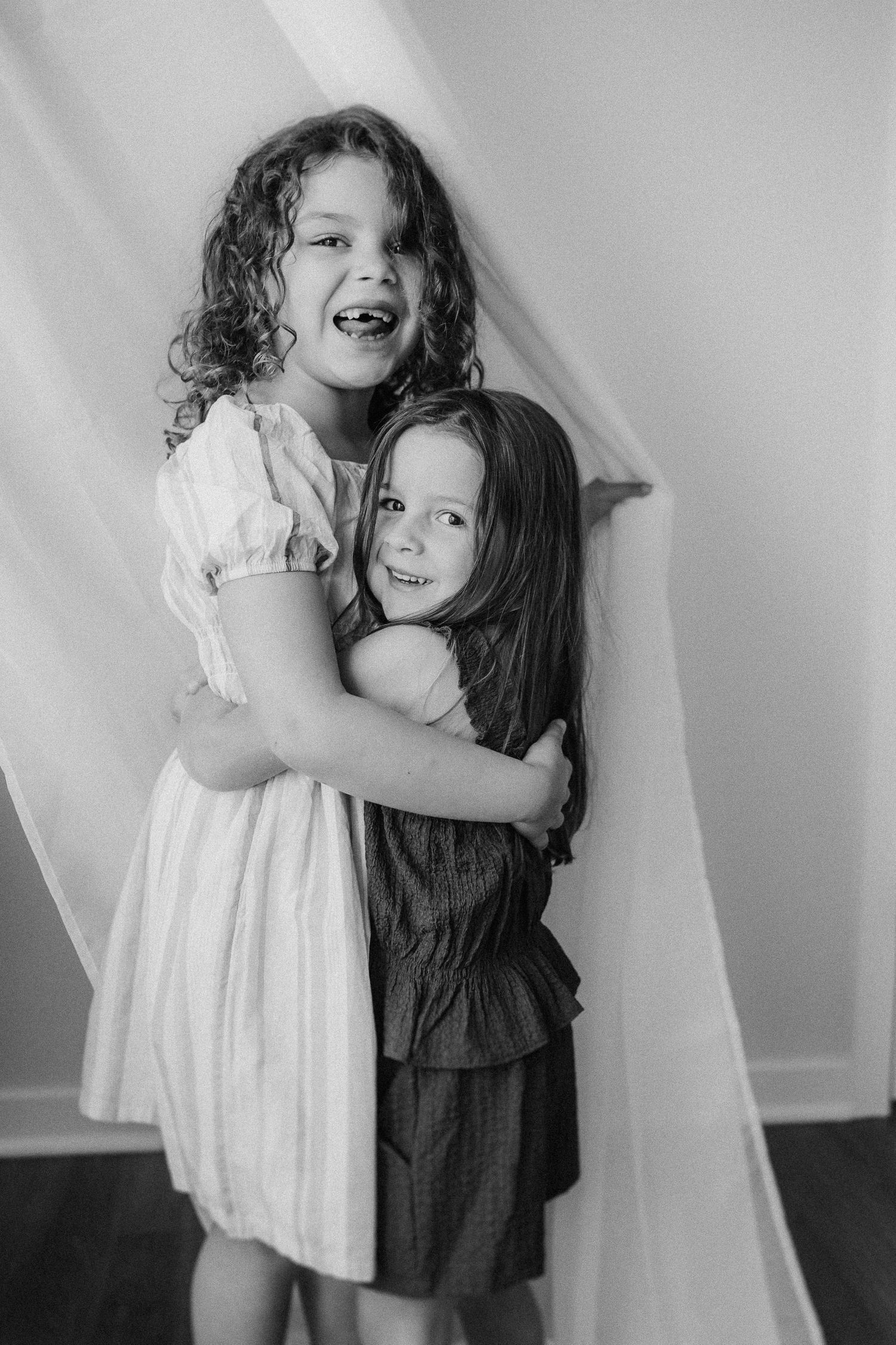 Two young sisters in contrasting dresses embrace and laugh together during a black and white portrait session.