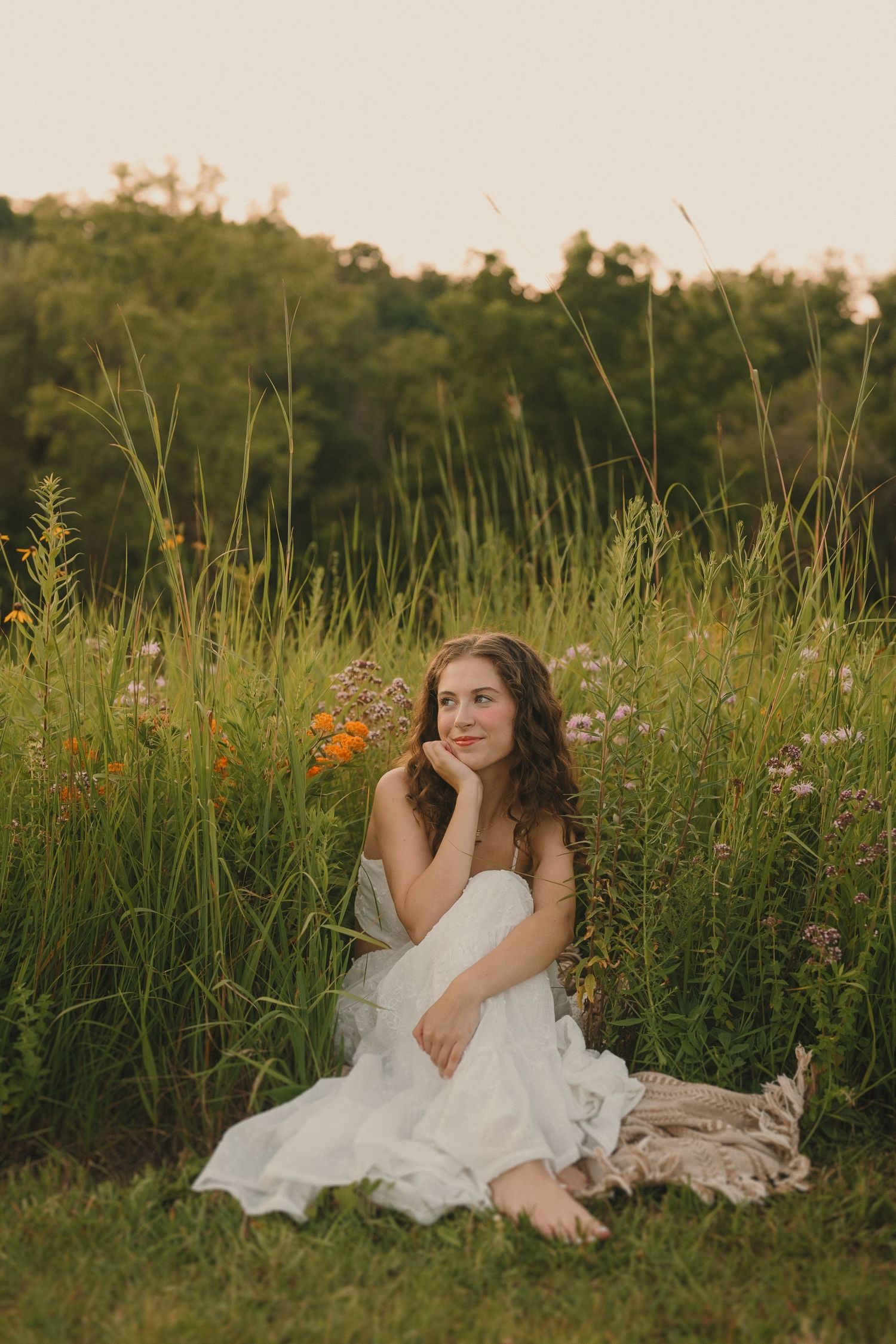 Someone in a white dress sits thoughtfully among wildflowers in a meadow at golden hour.