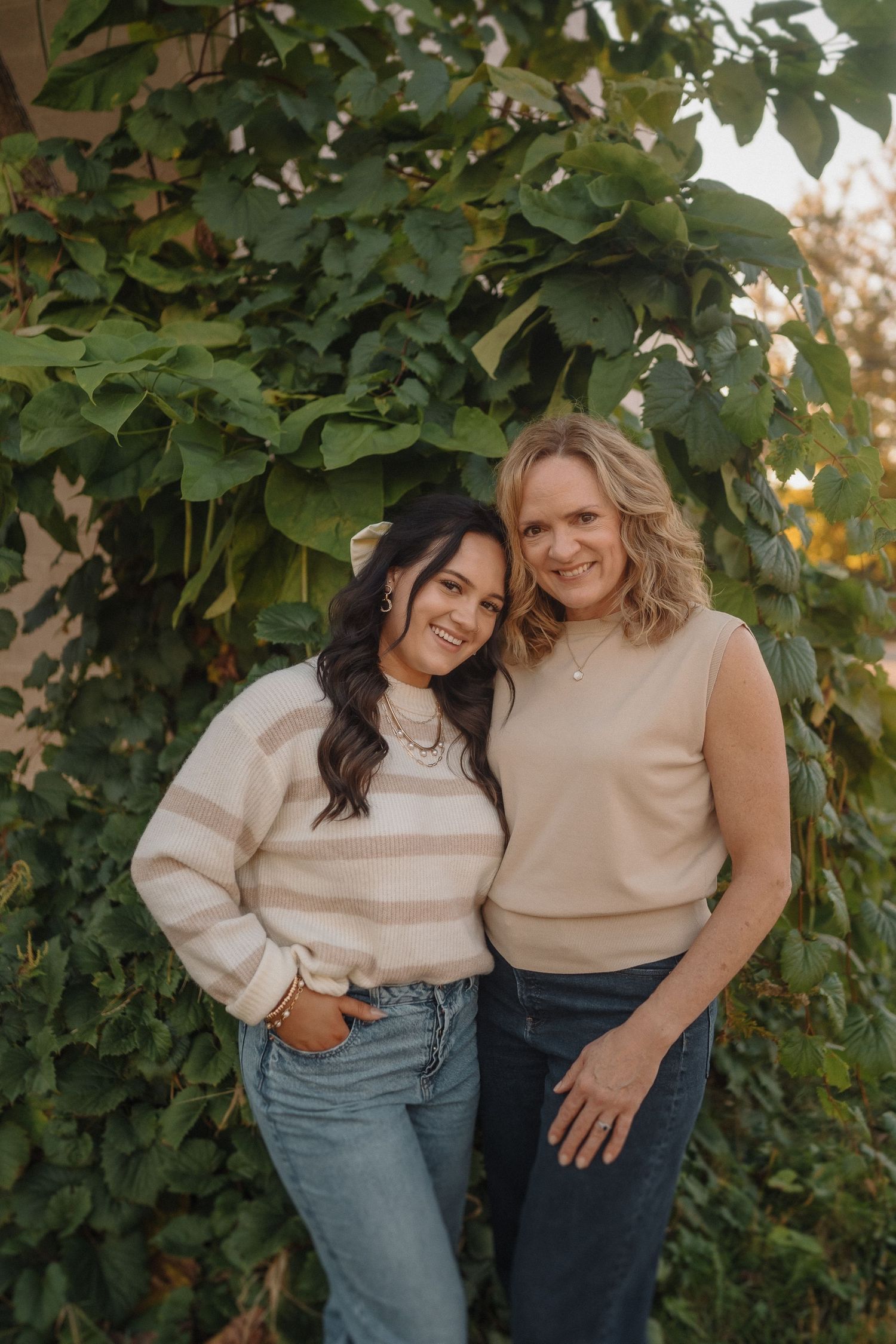 Two friends in neutral-colored sweaters and jeans pose together against lush green foliage during golden hour.