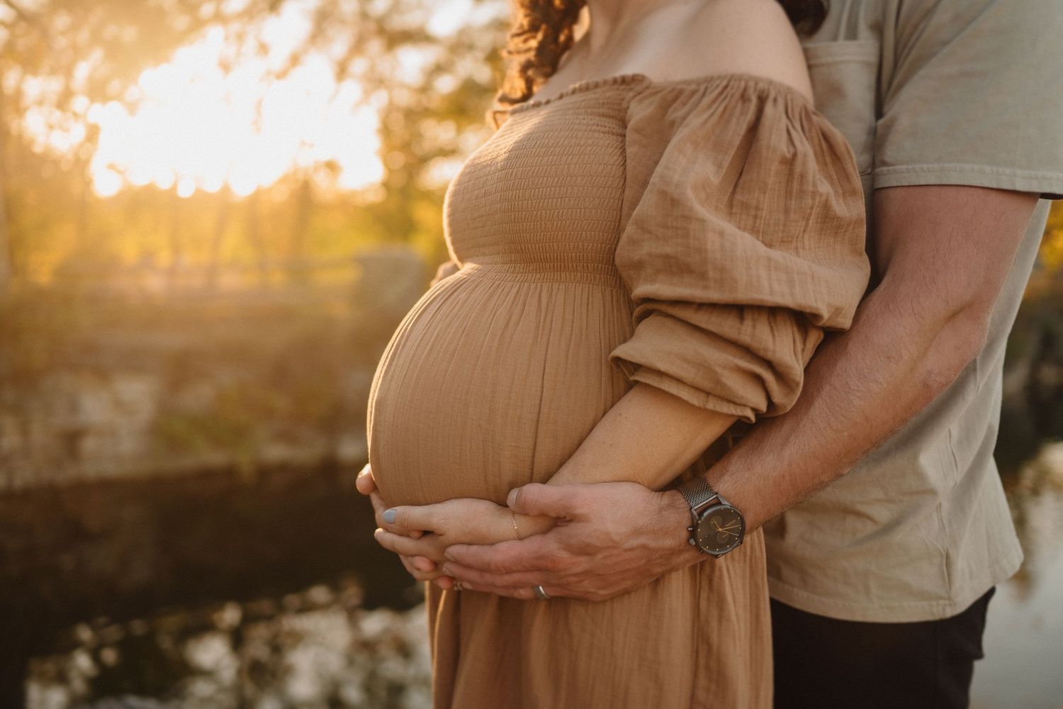 Sunset maternity photo showing hands embracing pregnant belly in brown dress by lake.