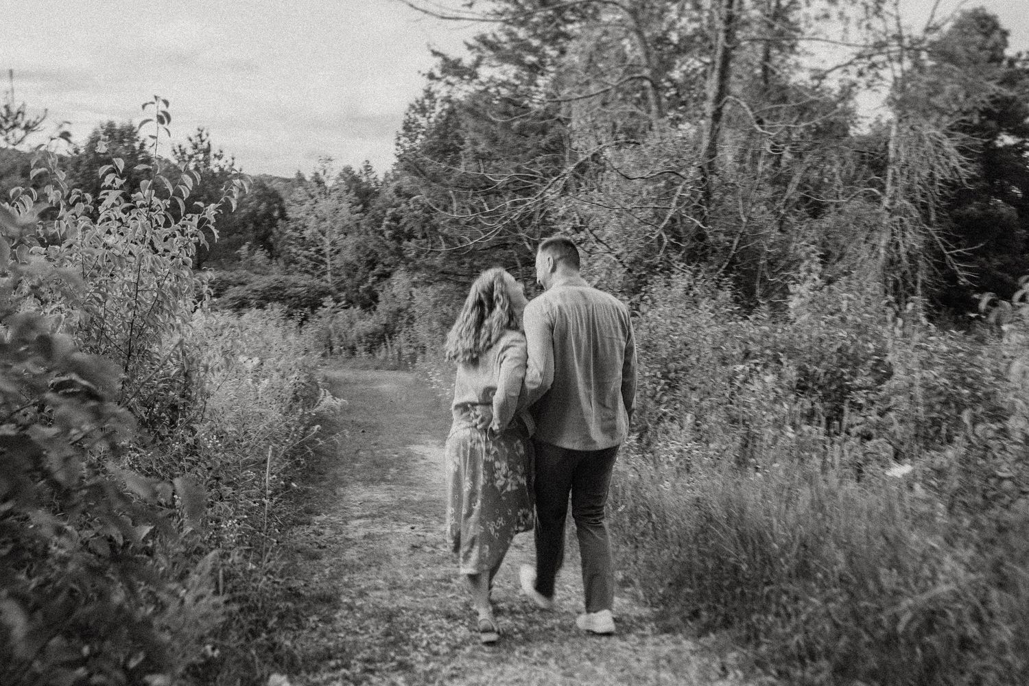 A couple walks away down a nature path surrounded by trees in black and white.
