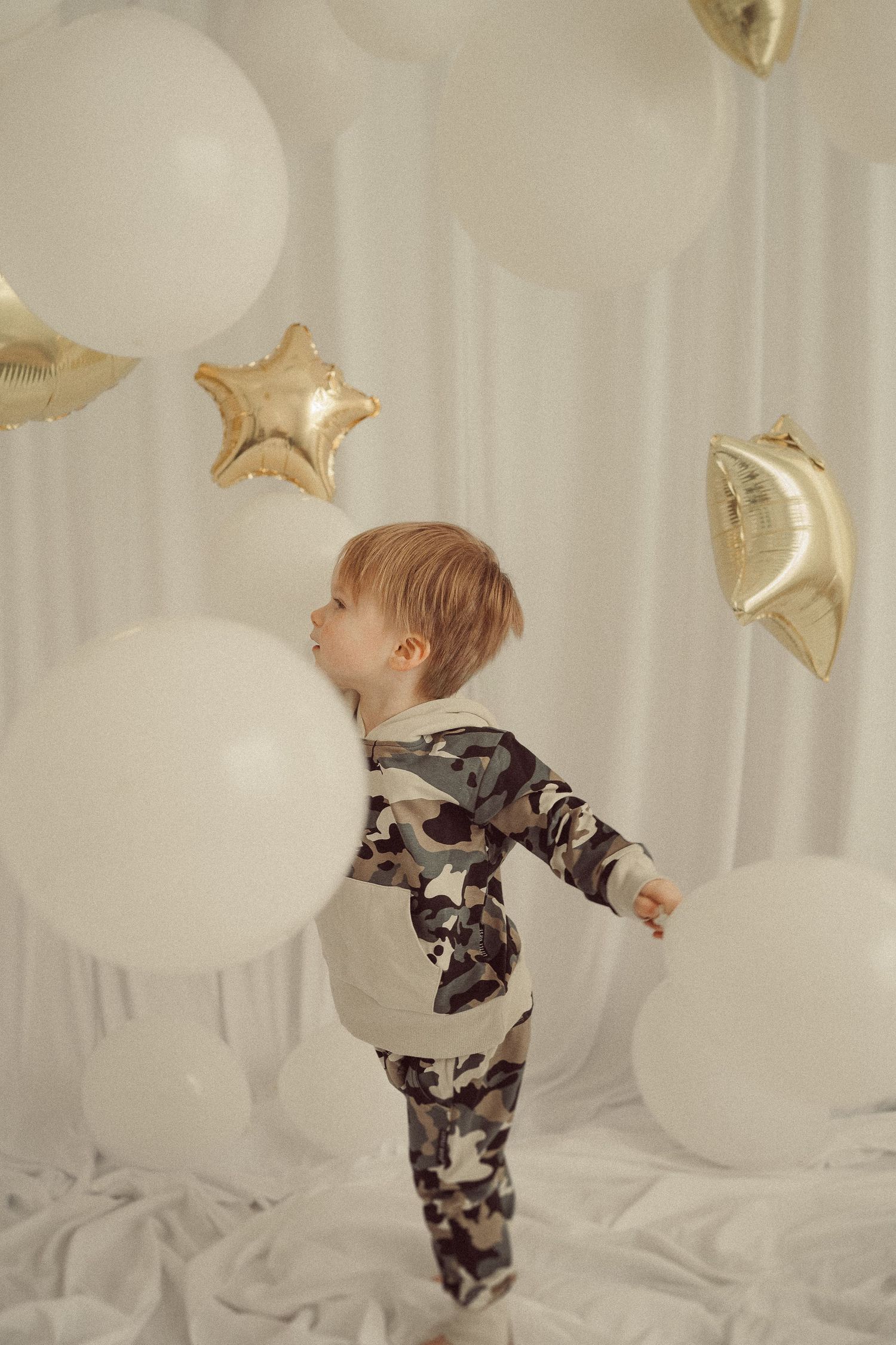 Child in camo outfit standing among white balloons and gold star decorations.