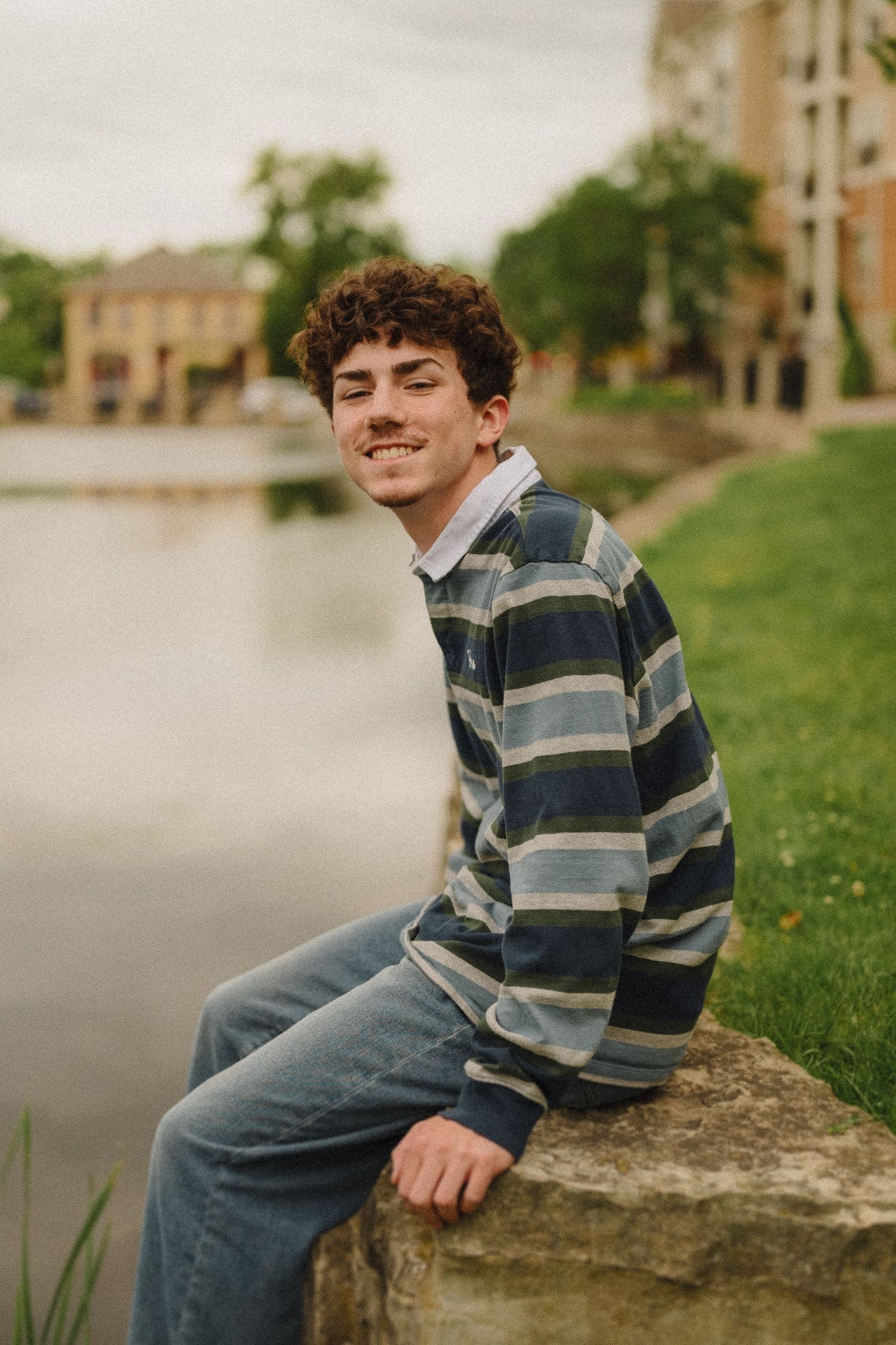 A young person in a striped sweater sits on a rock by a pond on an overcast day.