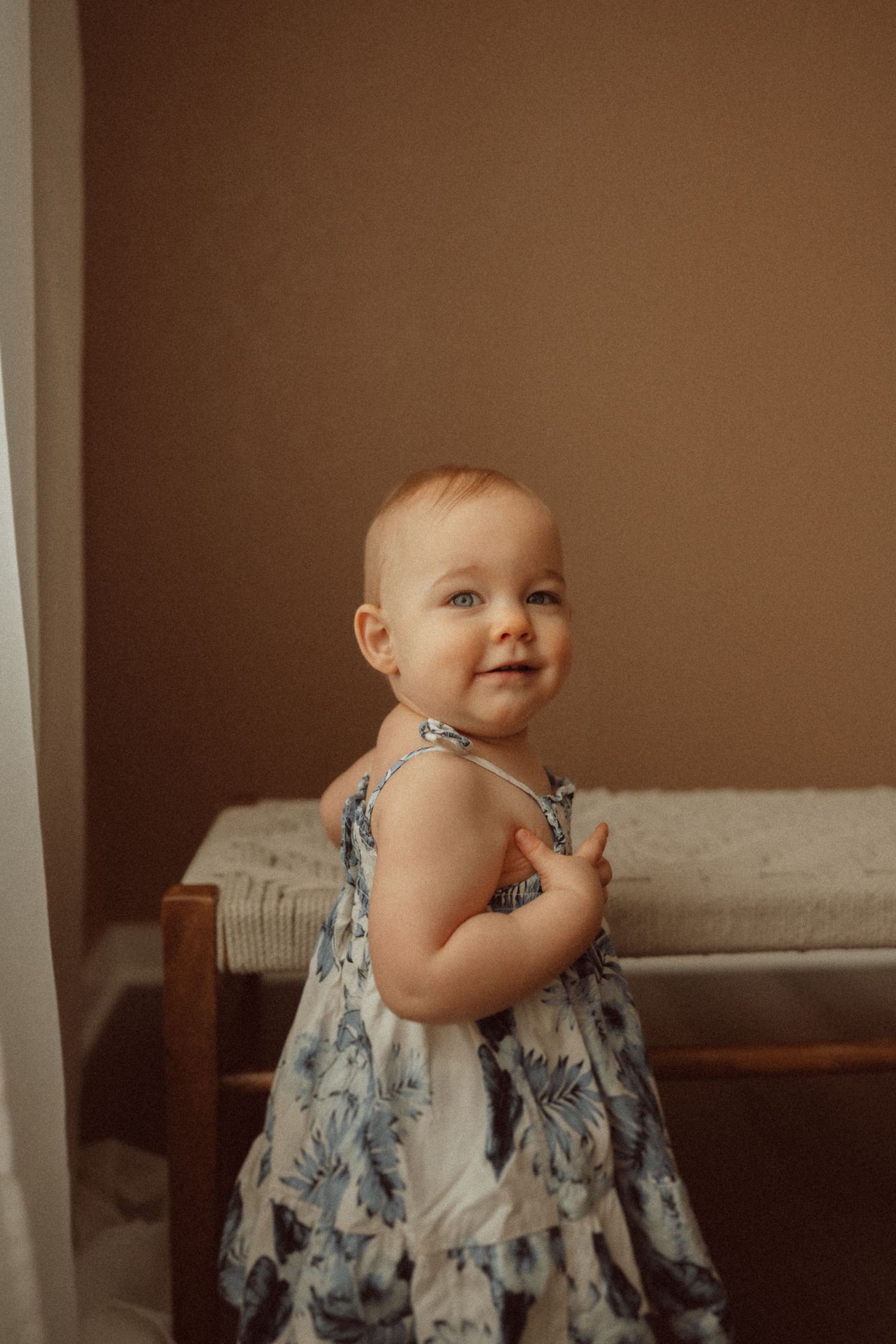 A small child in a blue floral dress poses near sheer curtains.