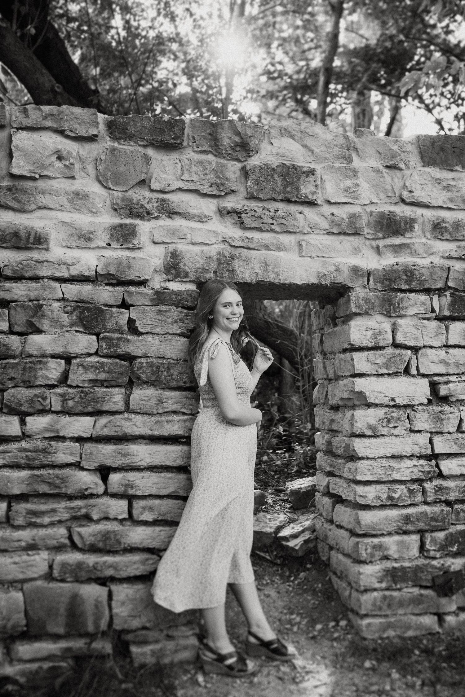 Black and white portrait of woman in dress posing beside stone wall archway.