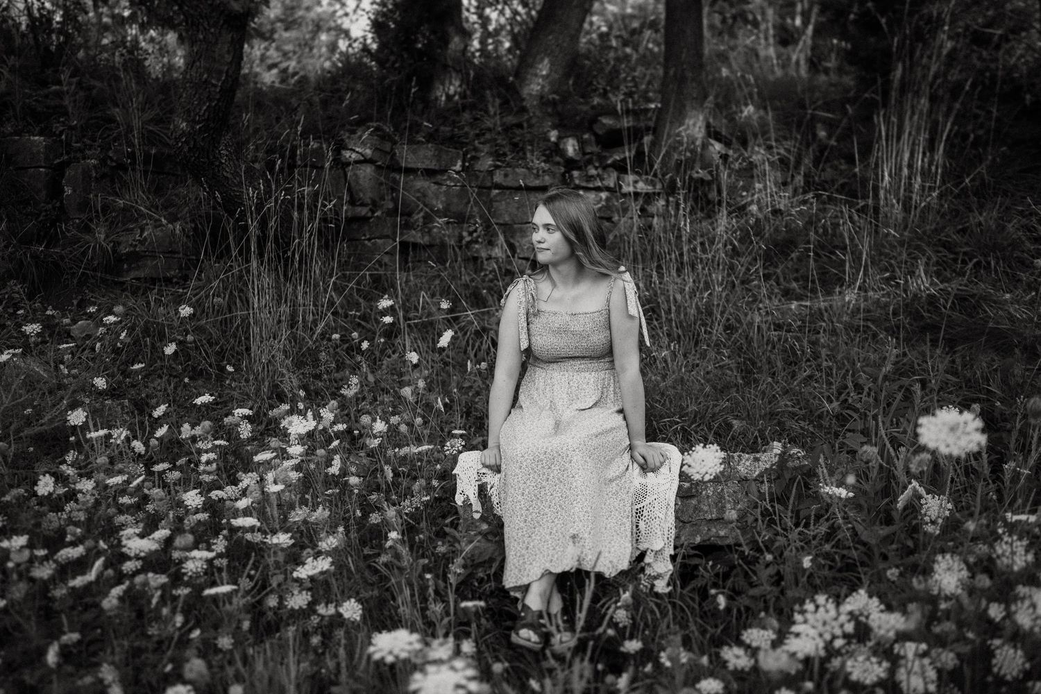 A black and white portrait amid blooming wildflowers against a rustic stone wall backdrop.
