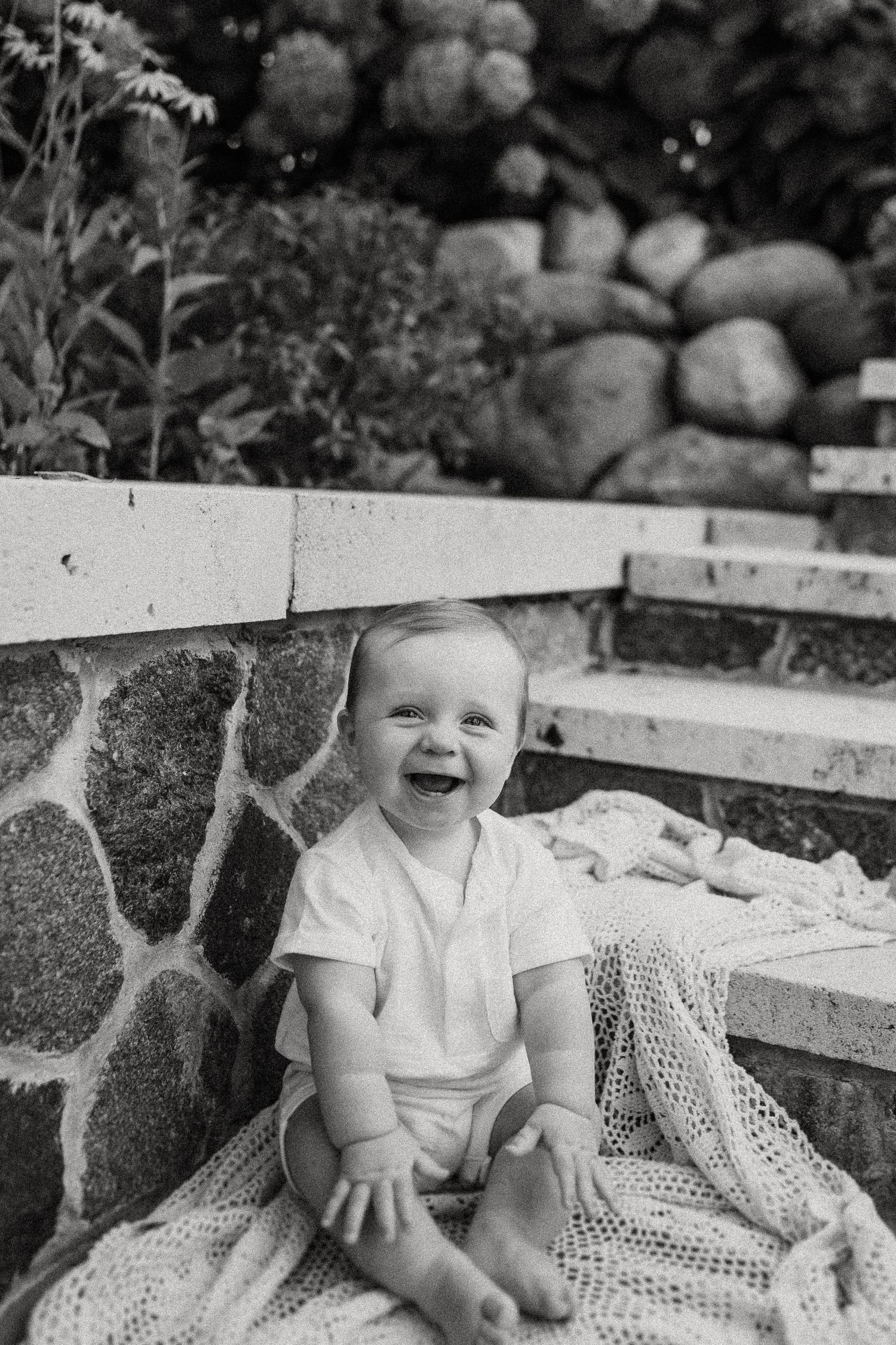 Black and white portrait of smiling baby sitting on lace blanket against stone steps.