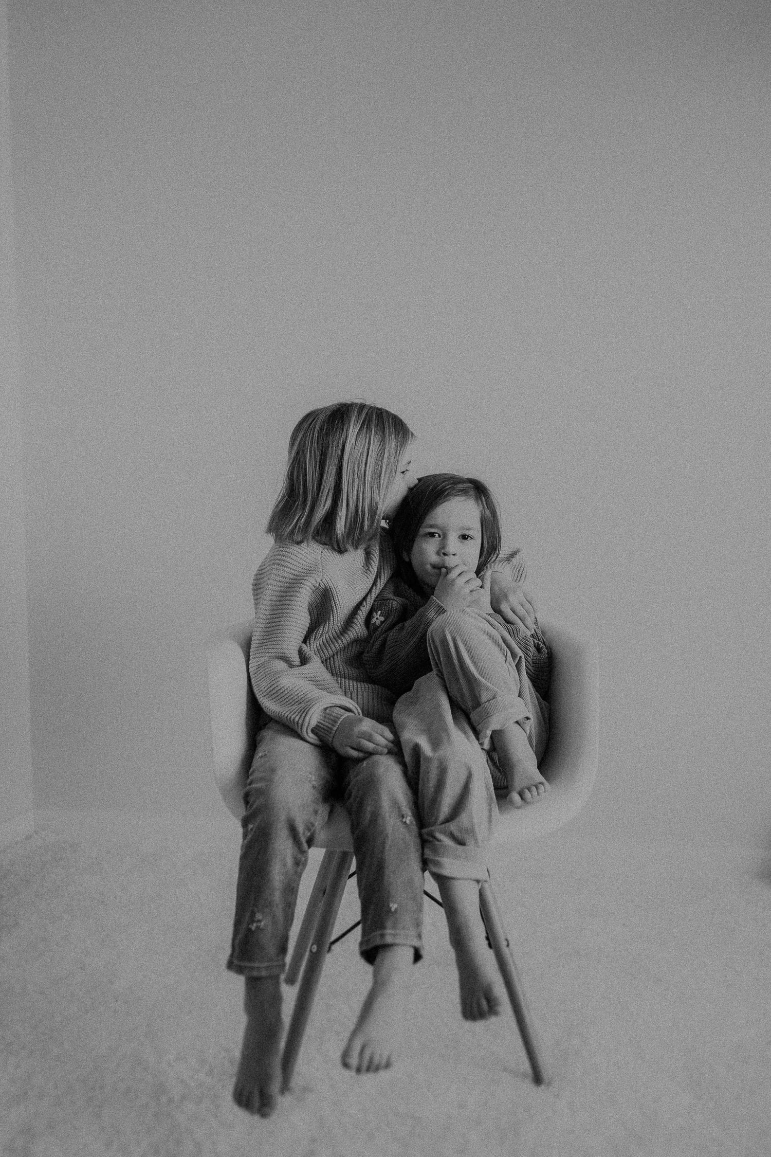 Two children sitting together on a modern chair in intimate black and white portrait.