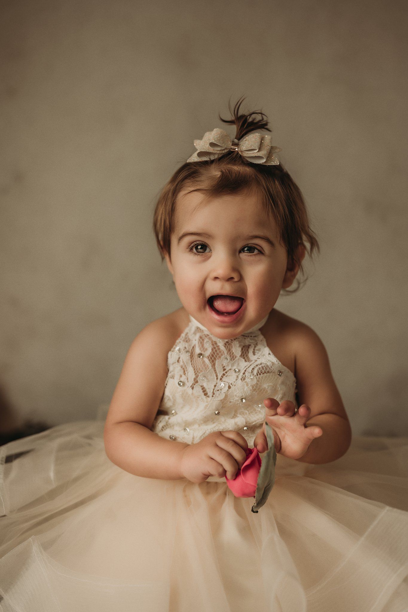A joyful small child wearing a cream tulle dress and bow hairpiece smiles brightly during a portrait session.