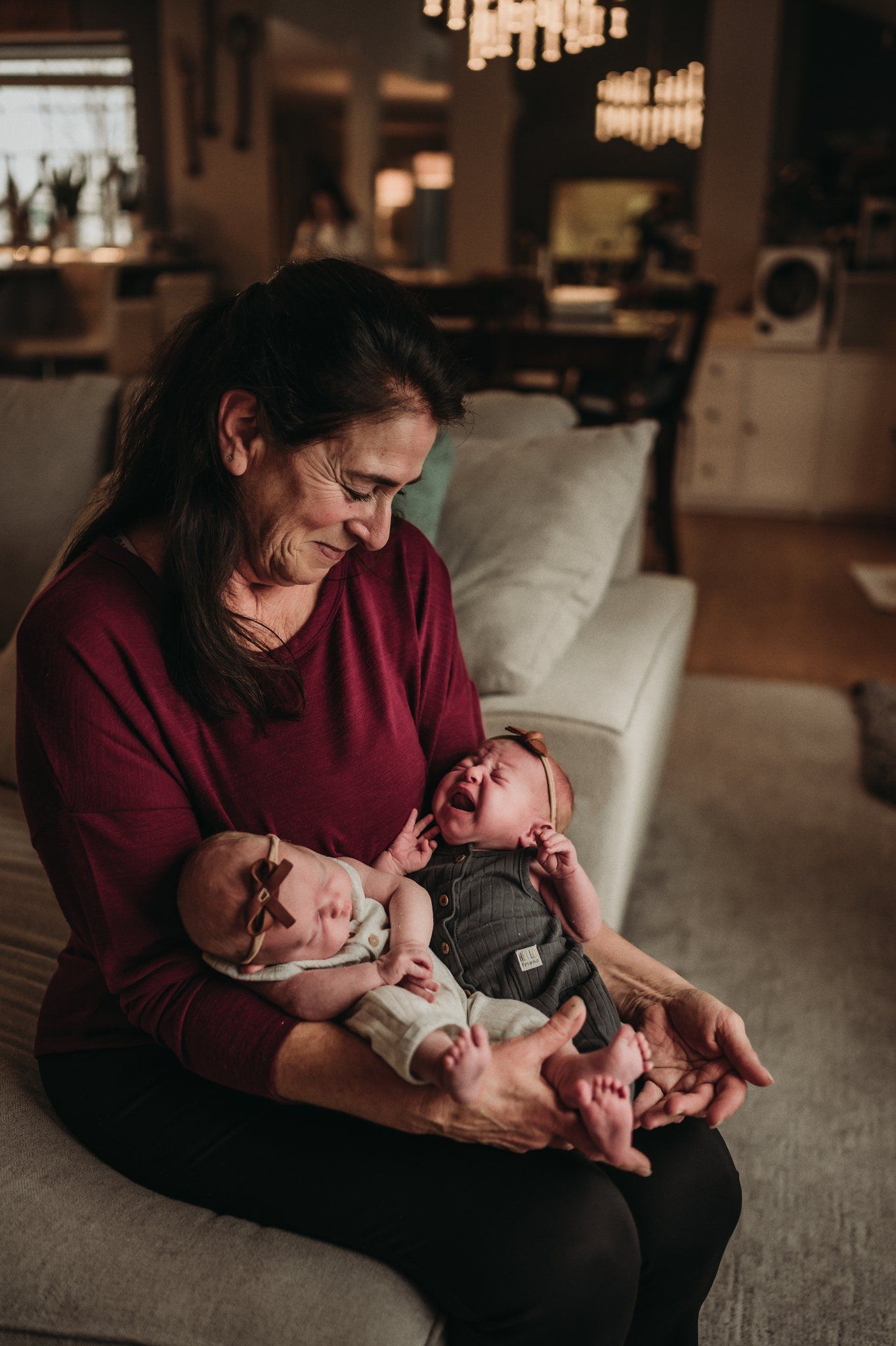 A cozy indoor moment of caregiver cuddling twin infants on a sofa under warm lighting.