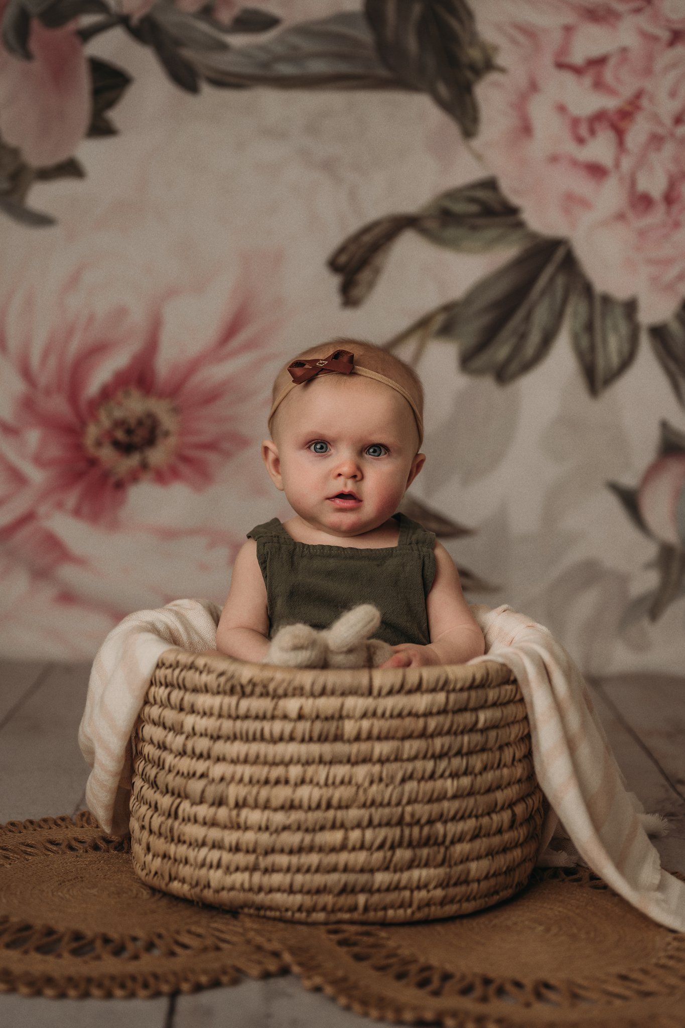 Baby seated in round woven basket against pink floral backdrop.