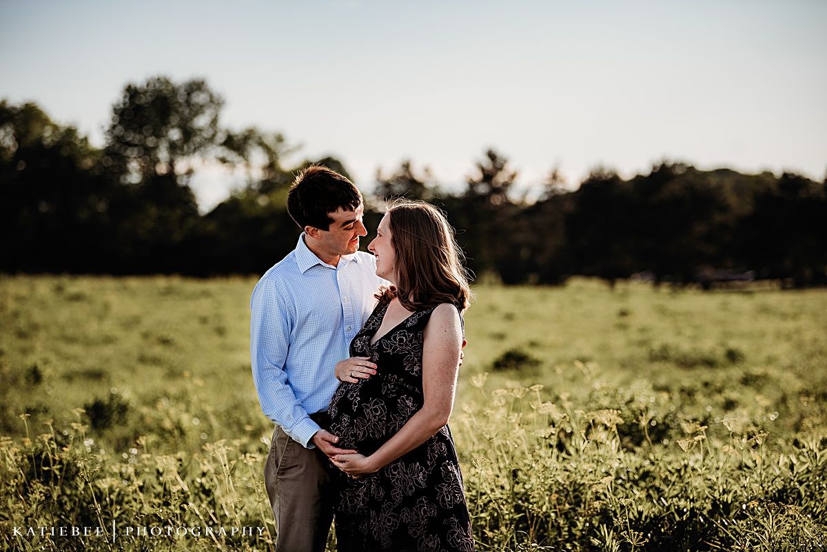 A couple stands together in a sunlit meadow during a maternity photo session.