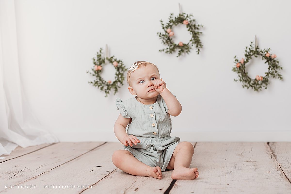 Baby sitting on wooden stool against white wall with floral wreaths.