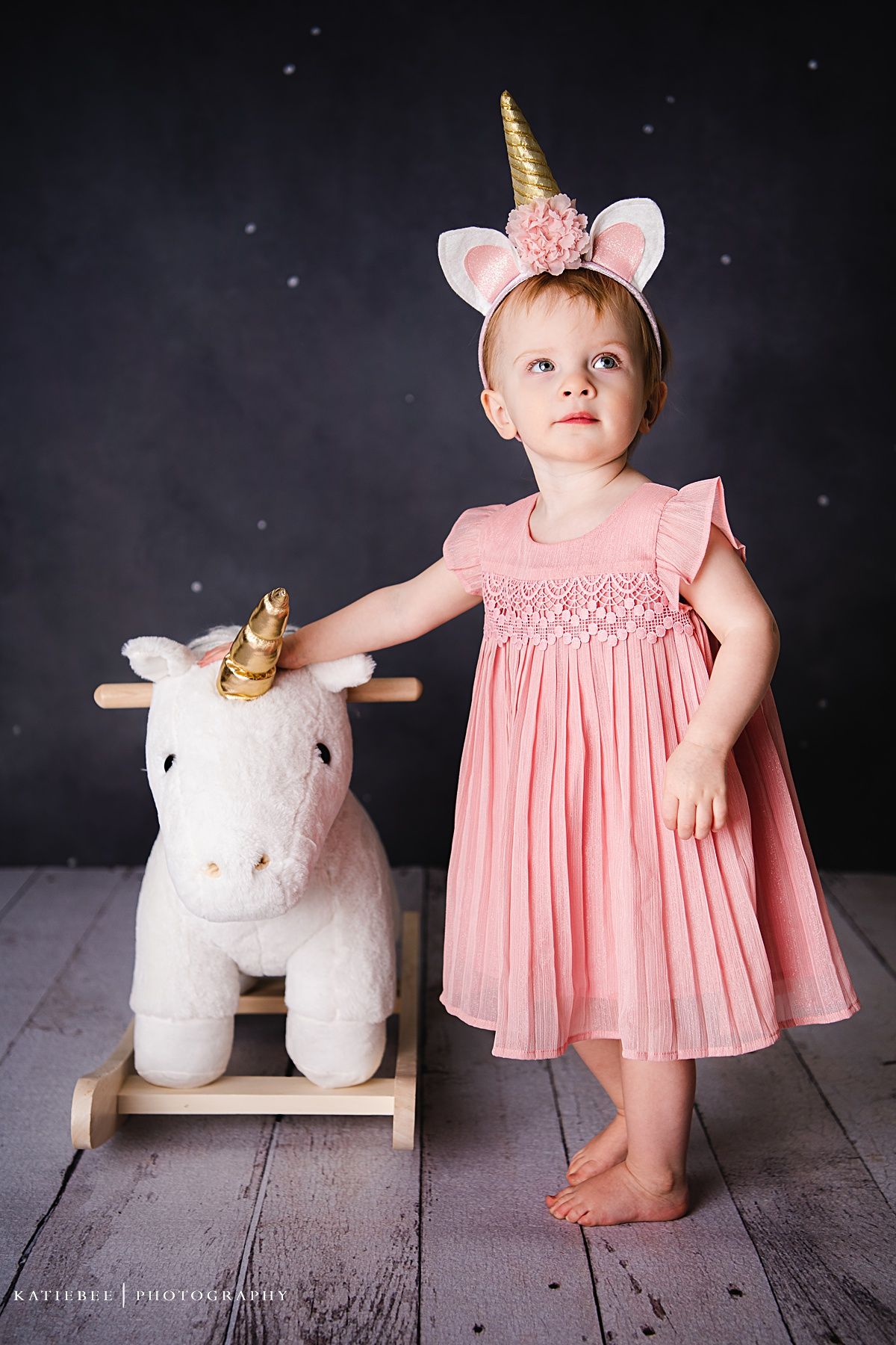 A small child in a pink dress and unicorn headband stands beside a white rocking unicorn toy.