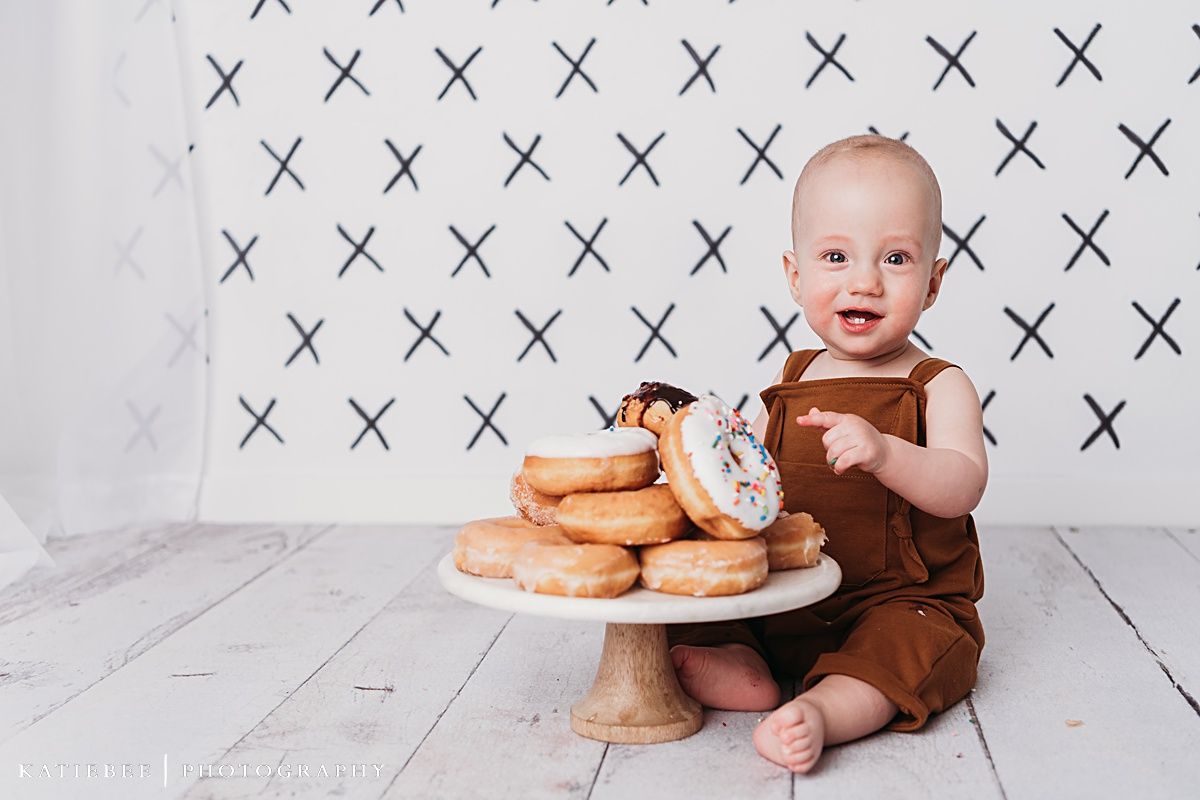 Baby in brown overalls sits next to plate of donuts on white wooden floor with X pattern backdrop.