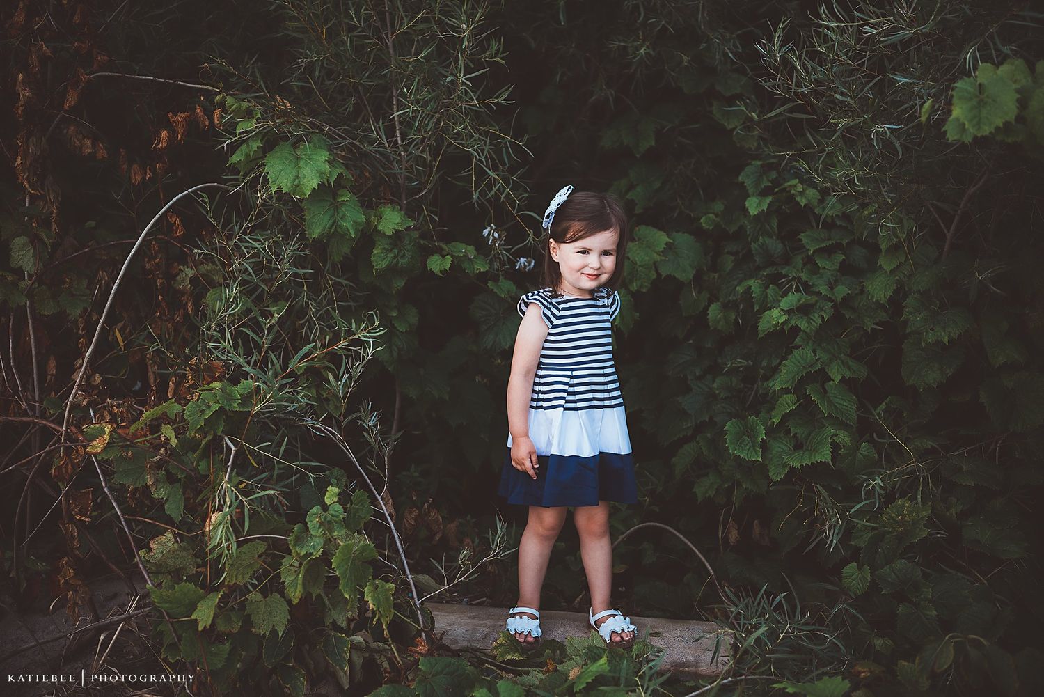 A young child in striped dress stands alone against a backdrop of green foliage.
