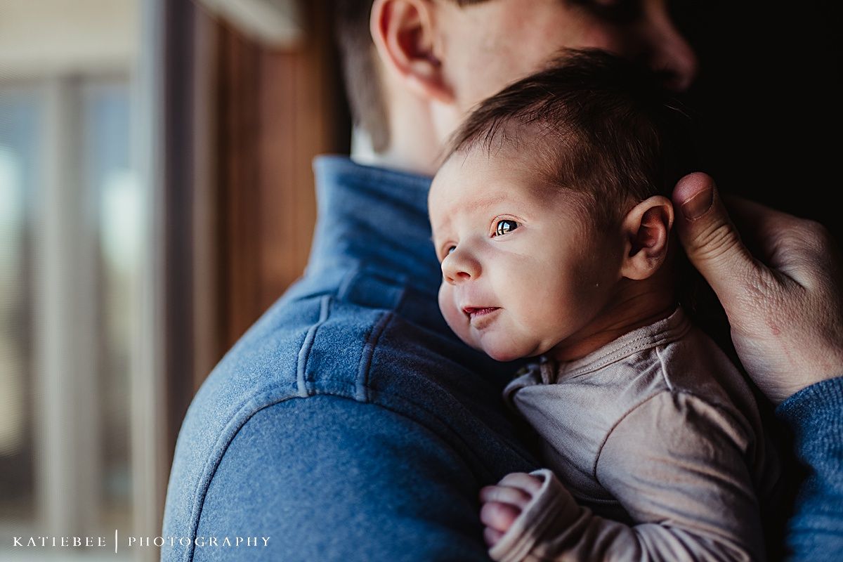 Side view of infant in beige outfit being held against blue clothing near window.