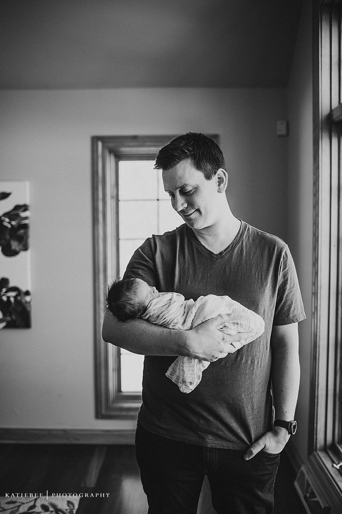 Black and white portrait of parent gazing lovingly at sleeping newborn by window.