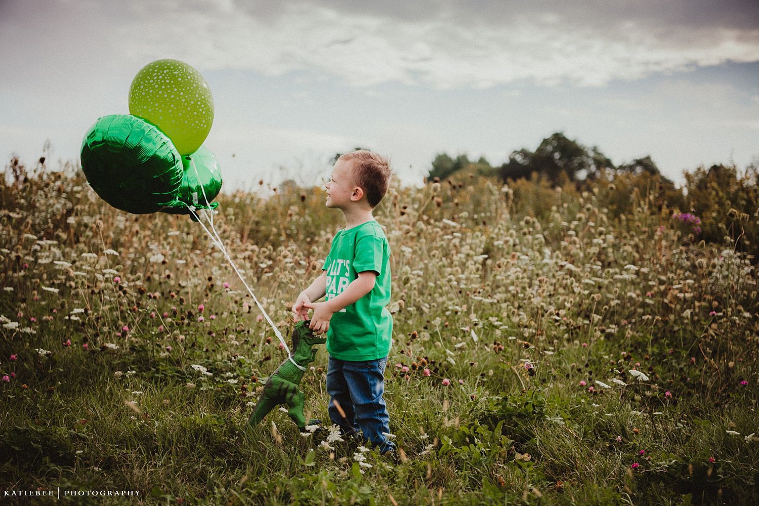 A child in green holds colorful balloons while standing in a wildflower field.