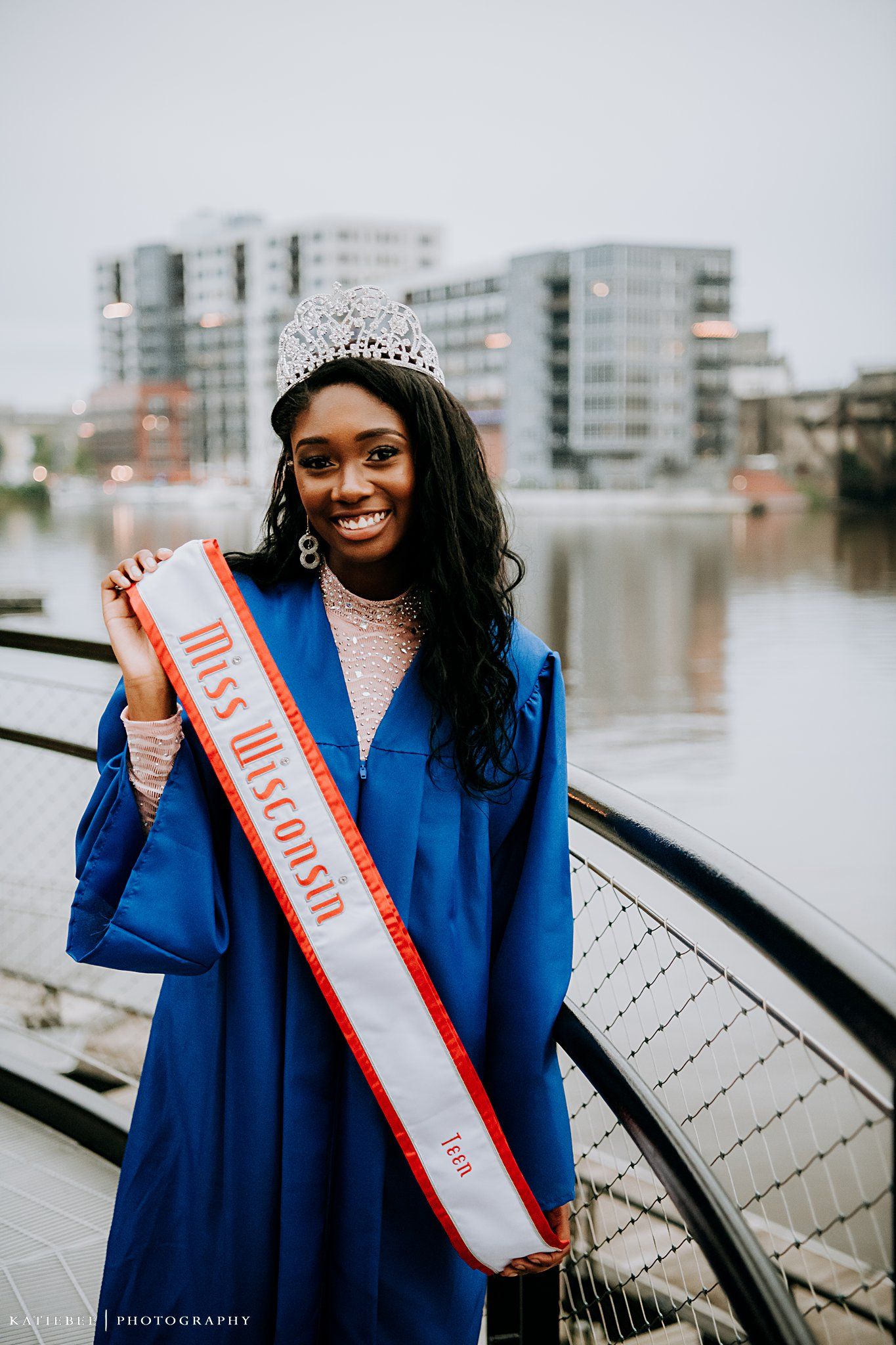 Graduate wearing tiara and sash poses in blue graduation gown by waterfront.