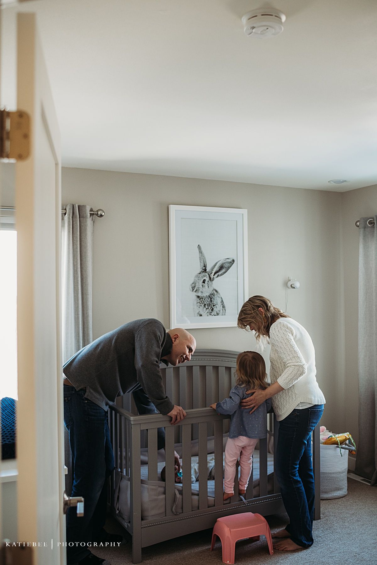 Family gathered around wooden crib in nursery with rabbit artwork on wall.