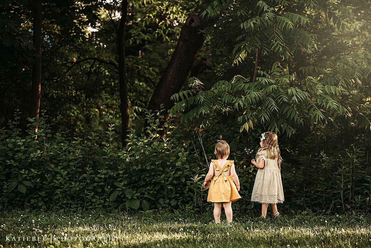 Two young children in light-colored dresses stand together at the edge of a wooded area.