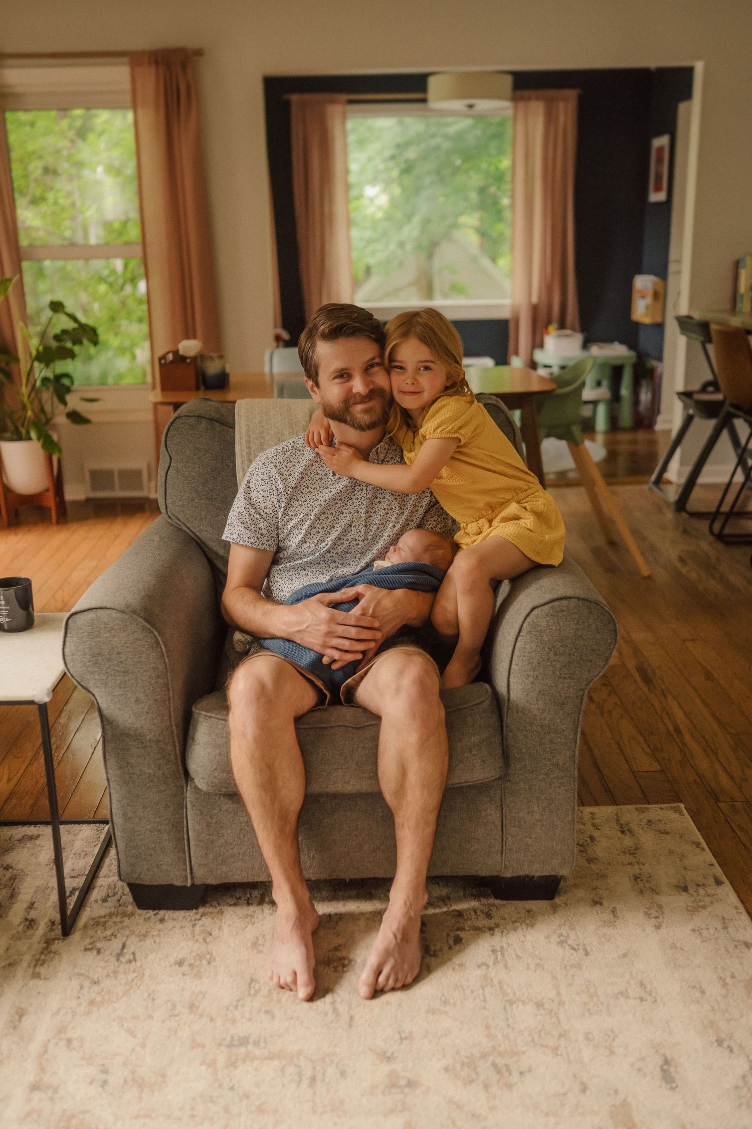 Family shares tender moment lounging on grey couch in sunlit living room.