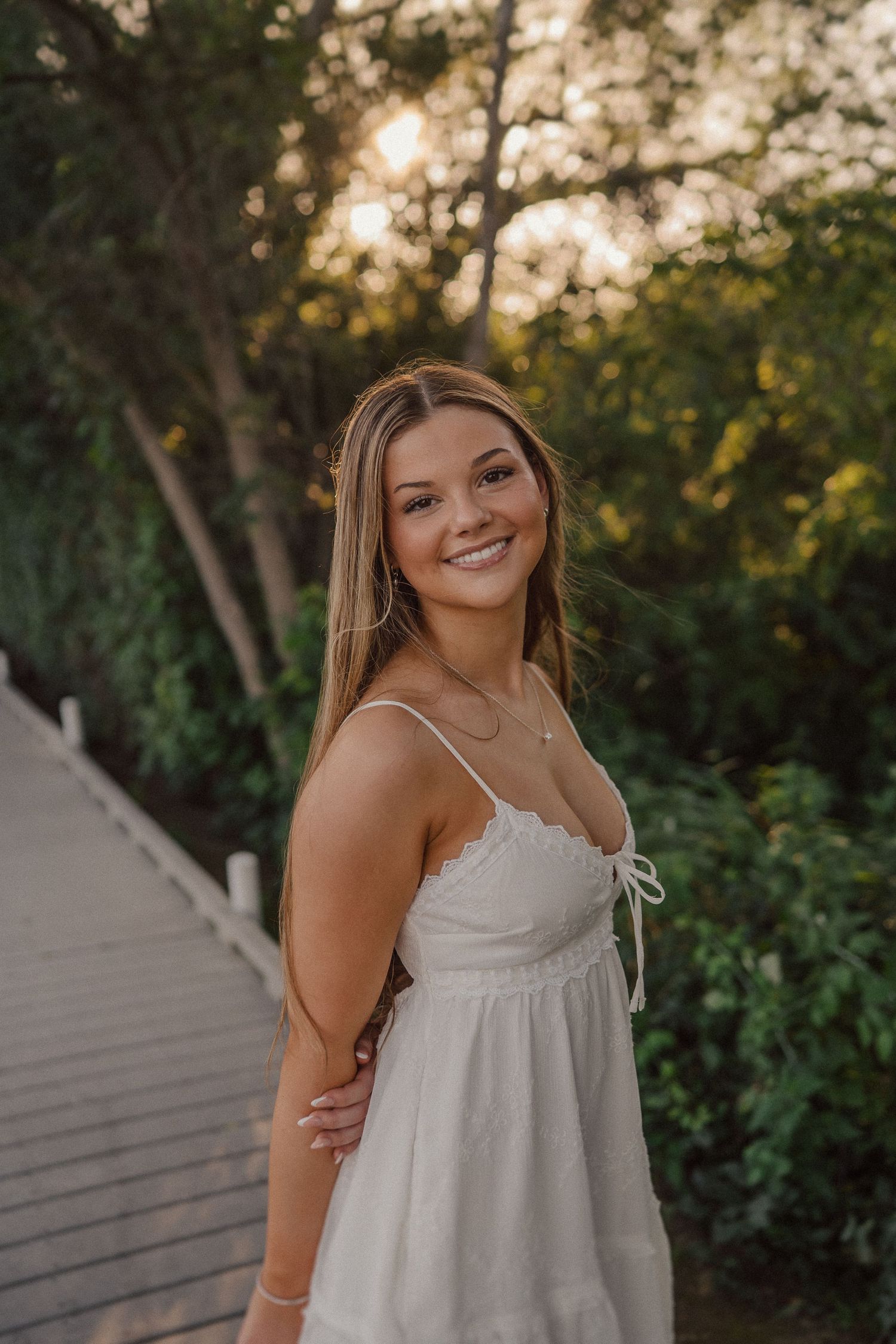 A woman in a white summer dress poses on a wooden walkway during golden hour.