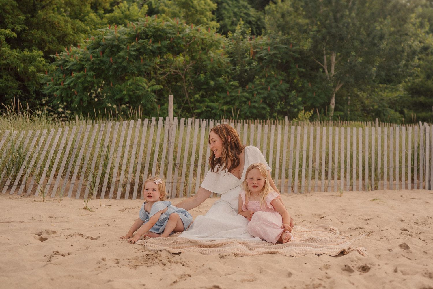 Family relaxing together on a sandy beach with wooden fence and greenery in the background.