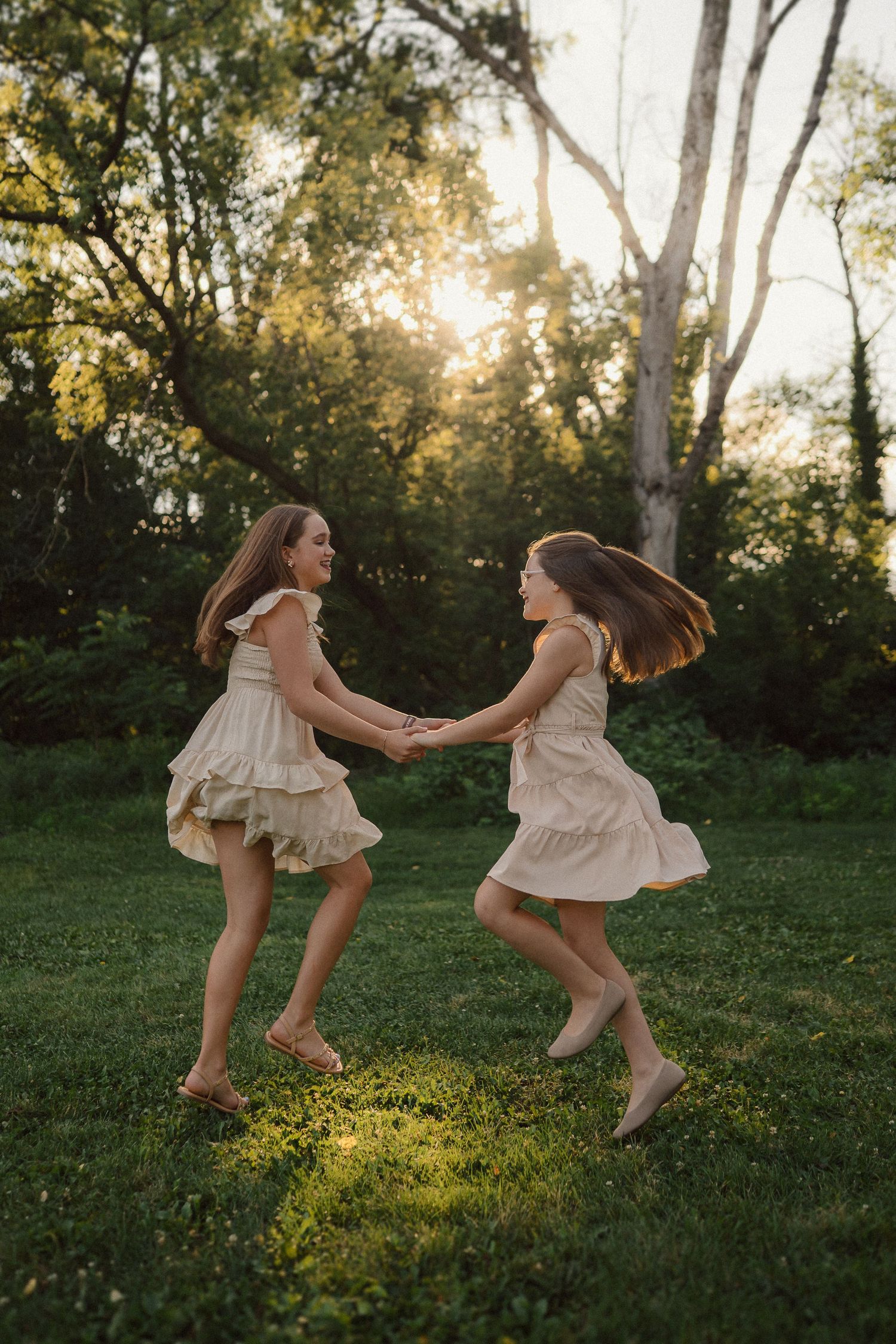 Two children in matching cream dresses dance joyfully in a sunlit garden at golden hour.
