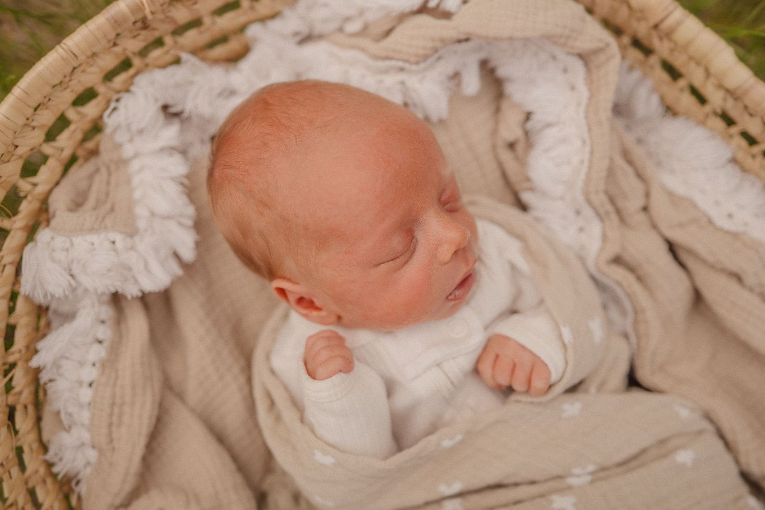 Sleeping newborn baby in white outfit nestled in textured basket with lace trim.