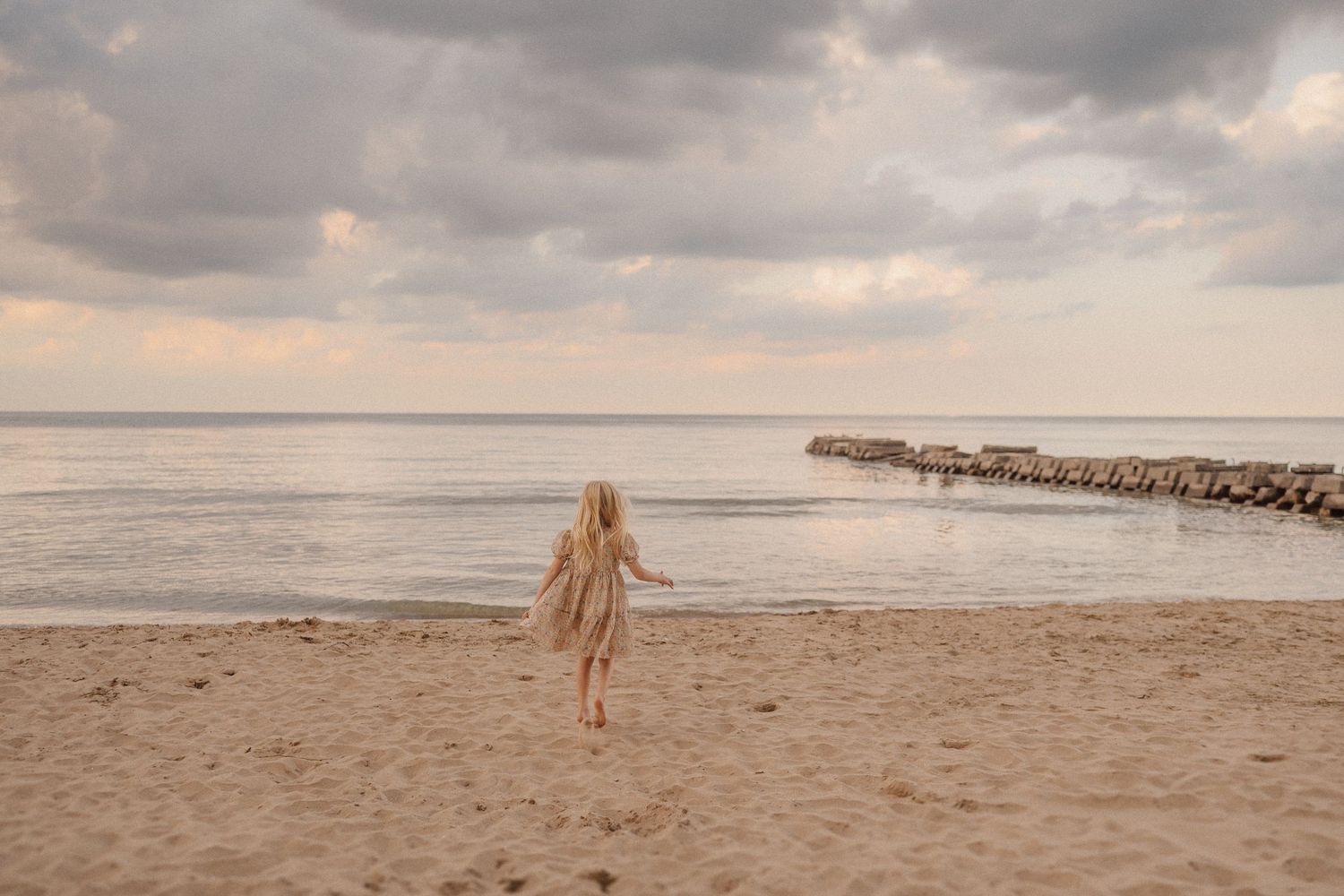 A lone figure walks on a sandy beach at sunset with a rocky jetty extending into the calm ocean under cloudy skies.