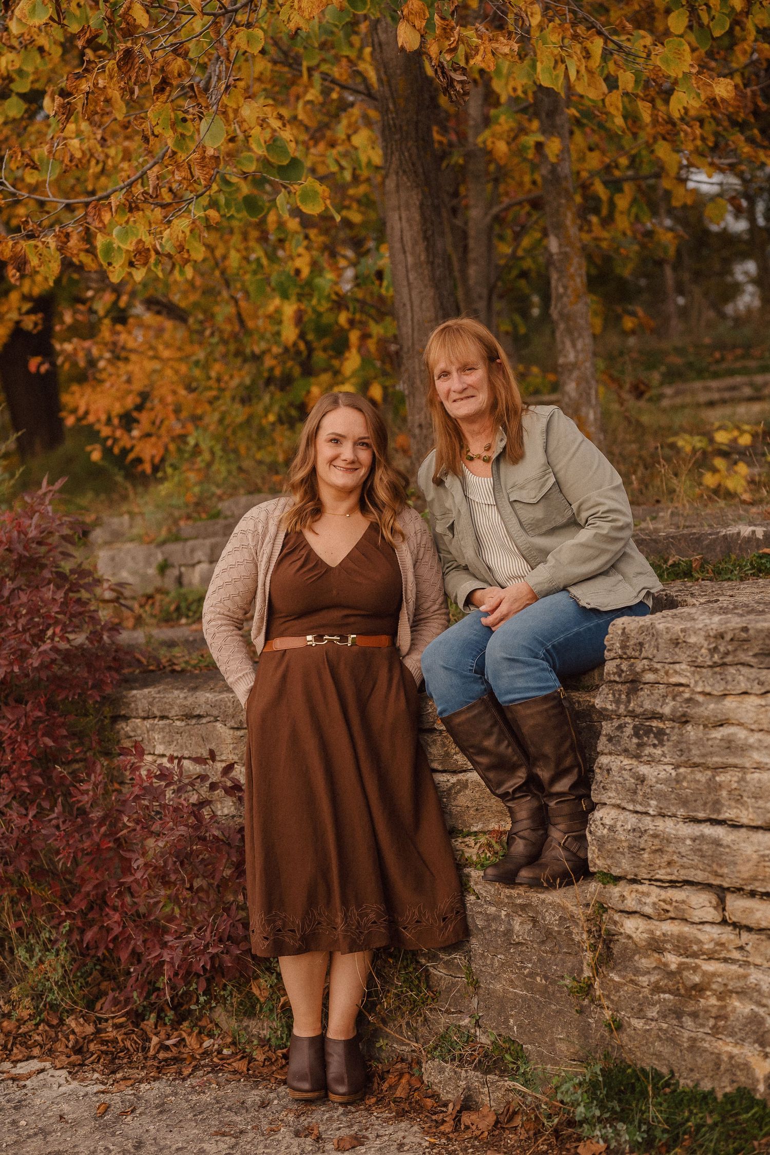 Two people sitting on stone wall with autumn trees displaying golden leaves in background.