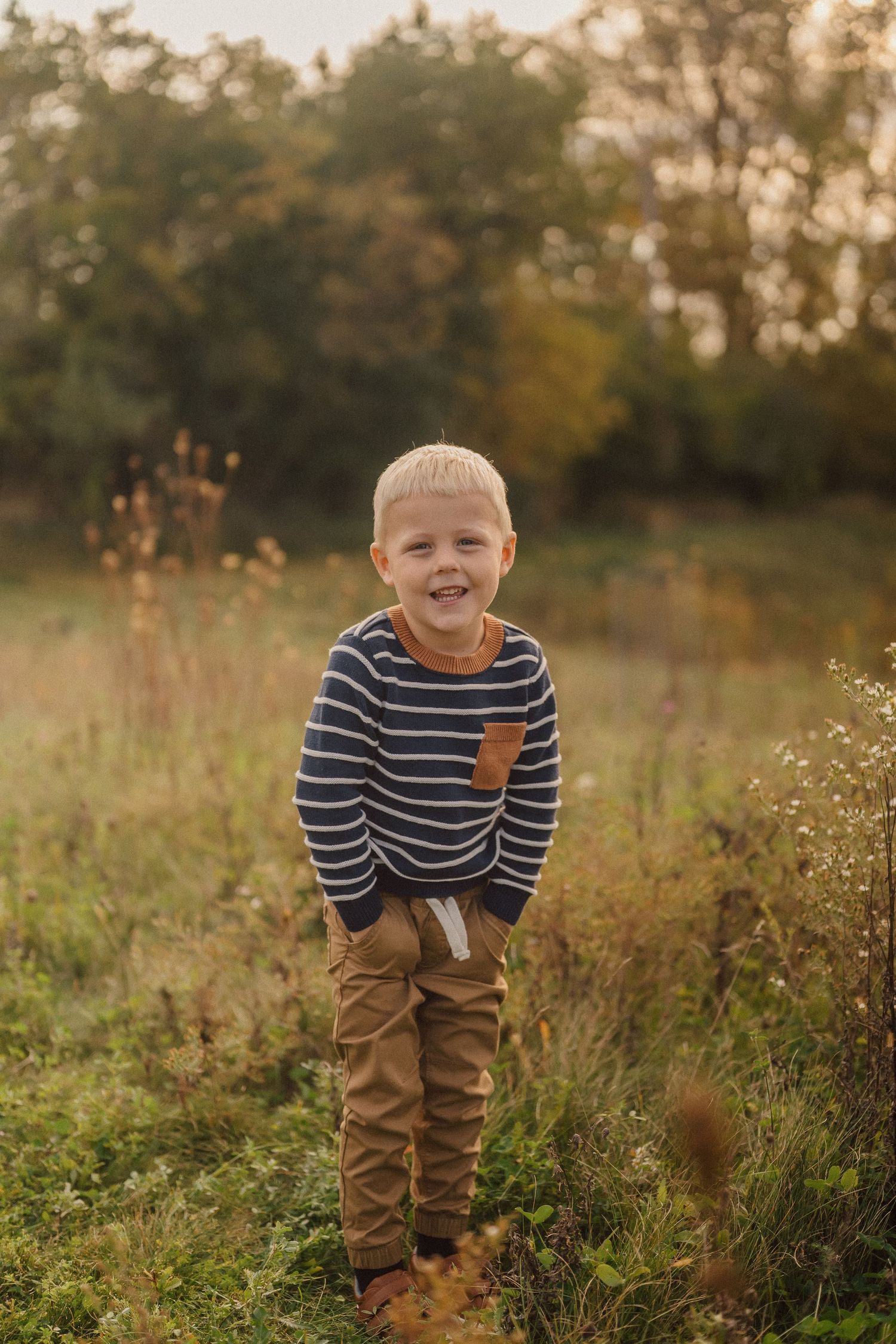 A child wearing a striped sweater and brown pants stands in a sunlit autumn meadow at golden hour.