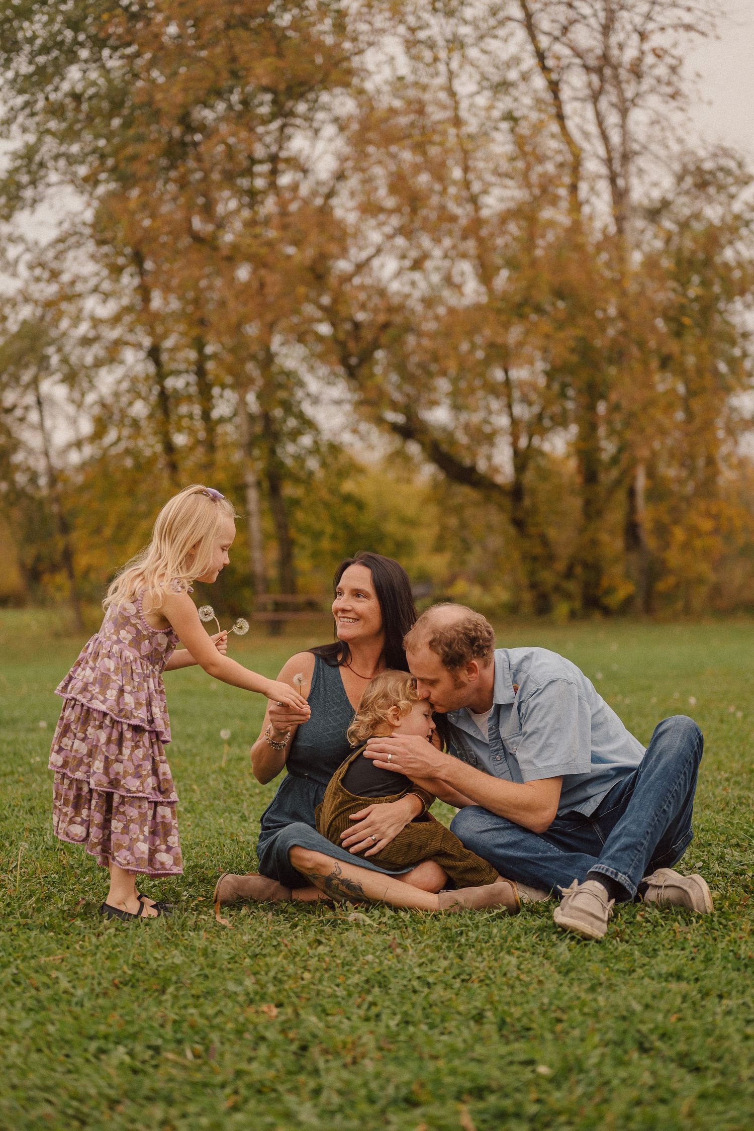 A family moment captured on green grass with autumn trees creating a warm backdrop.