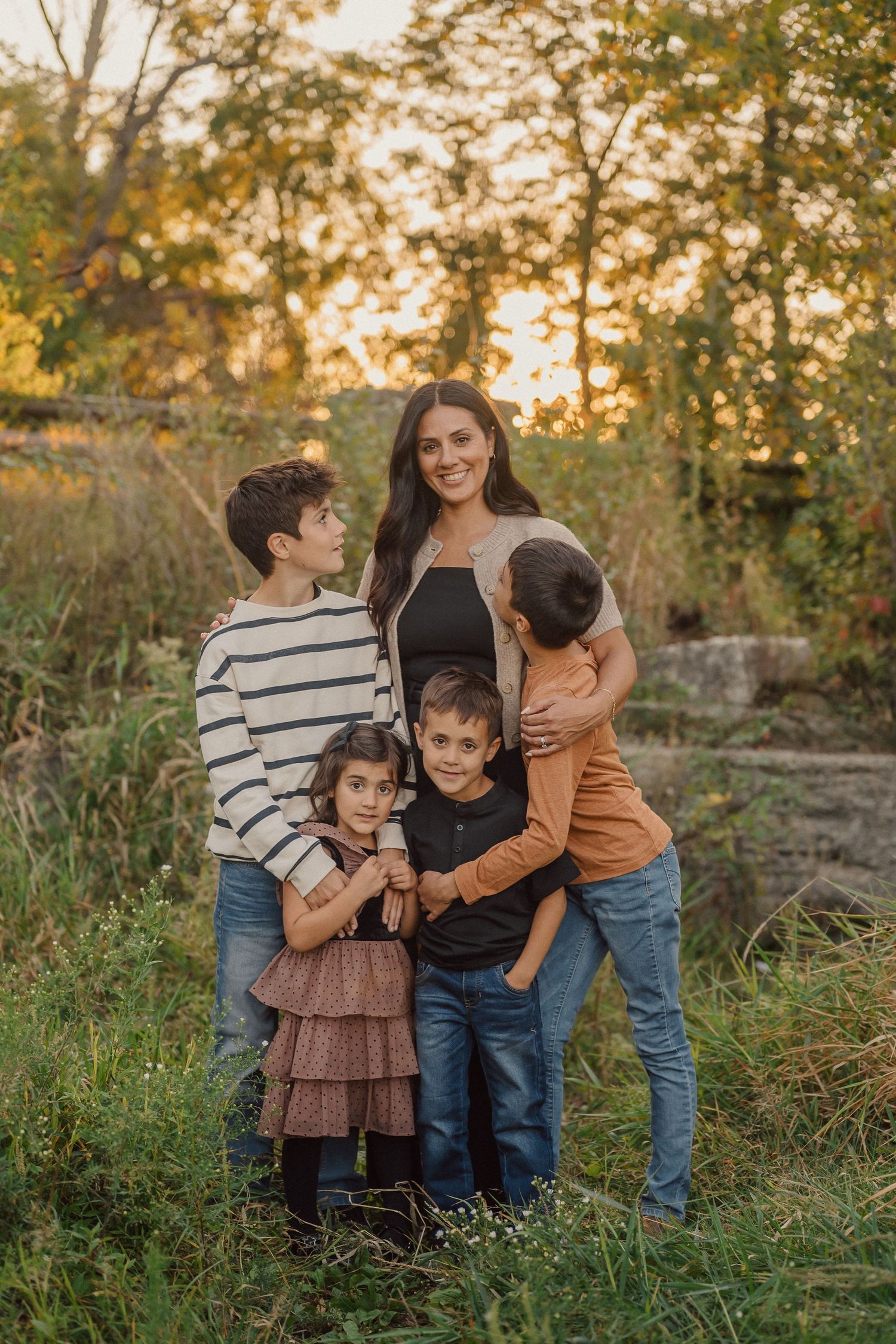 A family embraces outdoors during golden hour in a natural setting with tall grass and autumn foliage.