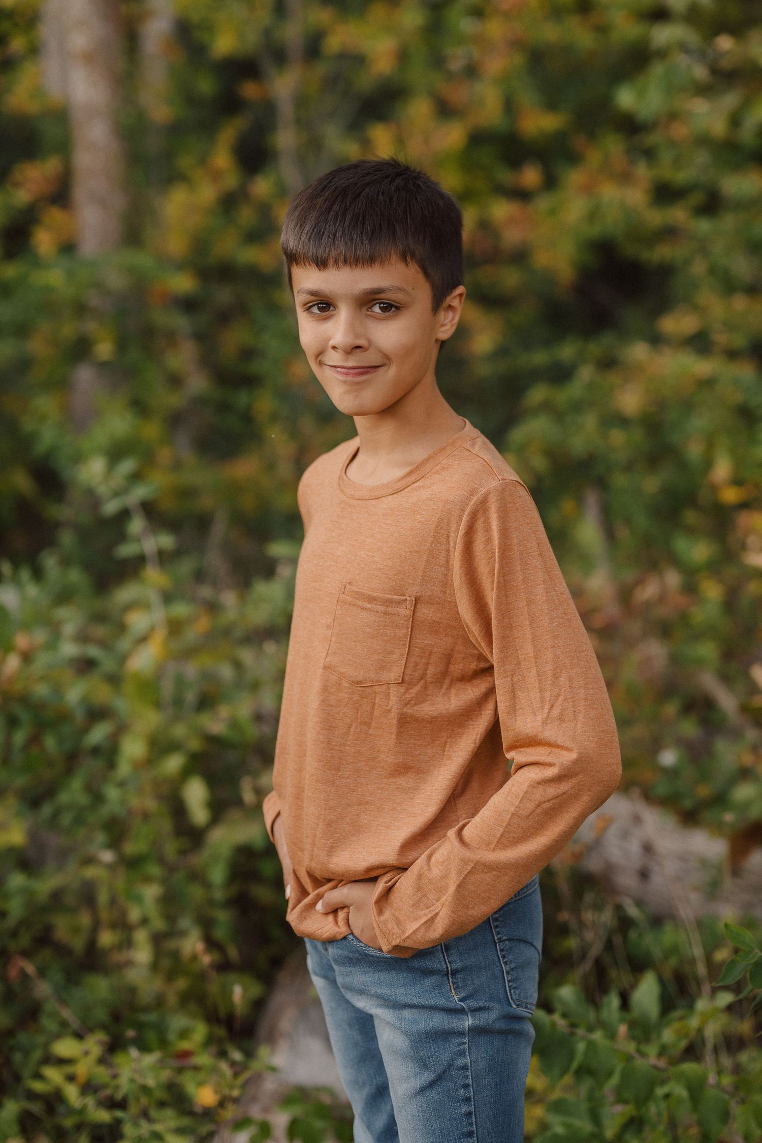 Young child in an orange long sleeve shirt and jeans poses outdoors with fall foliage in the background.